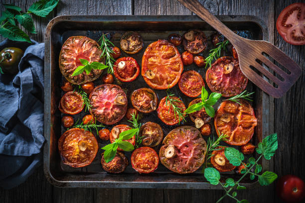 Roasted tomatoes cut varied in baking tray and ladle with basil and rosemary on wood and tomato plant leaves