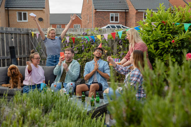 Group of friends having a garden party outdoors in the North East of England. They are sitting together taking pictures and singing with a cake on an outdoor patio.