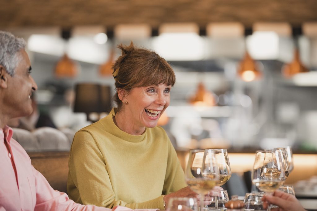 Close up shot of a mature woman laughing with friends at a restaurant meal. They are drinking white wine.